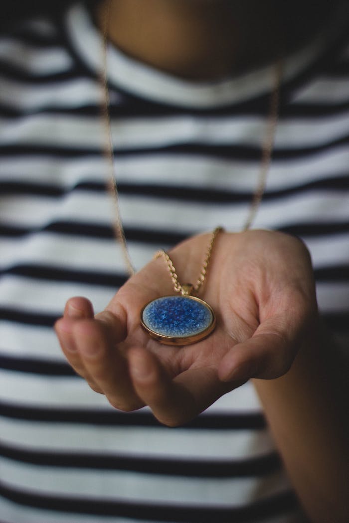 why-choose-us Close-up of a hand holding a blue pendant necklace against a striped shirt.